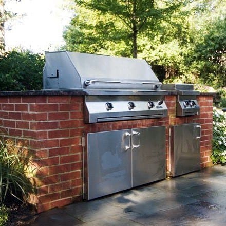 Built-in grill with stainless steel finish, set amid brick surround on a patio embraced by lush greenery