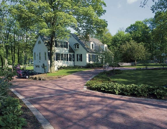 Red brick paver driveway with curved edging leading to a white house surrounded by trees.