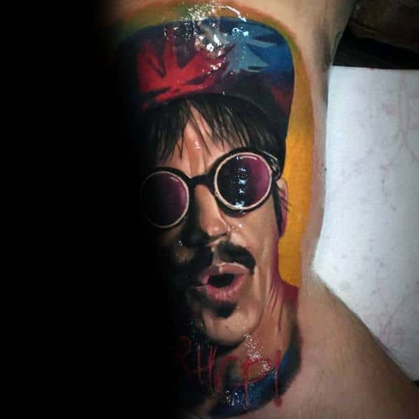 Realistic tattoo featuring a vibrant portrait of a singer wearing round sunglasses and a colorful cap with the Red Hot Chili Peppers logo.