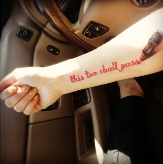A "This Too Shall Pass" tattoo on a person's forearm, with red cursive letters, alongside a lipstick tube.