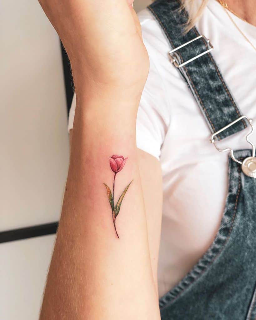A small, delicate pink tulip tattoo on the forearm, featuring fine details, a thin stem, and soft green leaves for a realistic floral design.