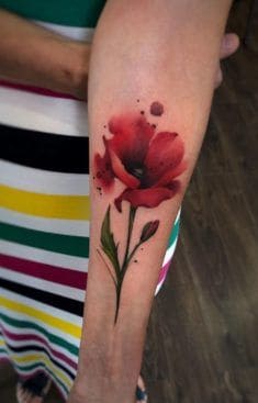 A vibrant watercolor-style red flower tattoo on the forearm, featuring bold petals, a green stem, and artistic ink splashes for a dynamic effect.