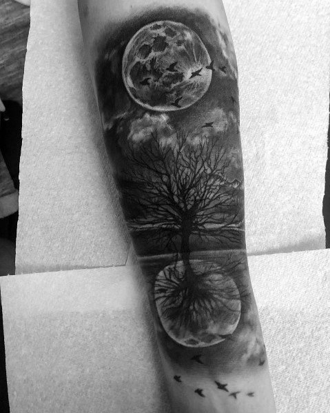 Tattoo of a full moon and leafless tree reflected in a lake, with birds flying across a cloudy sky, creating a mystical and surreal design.