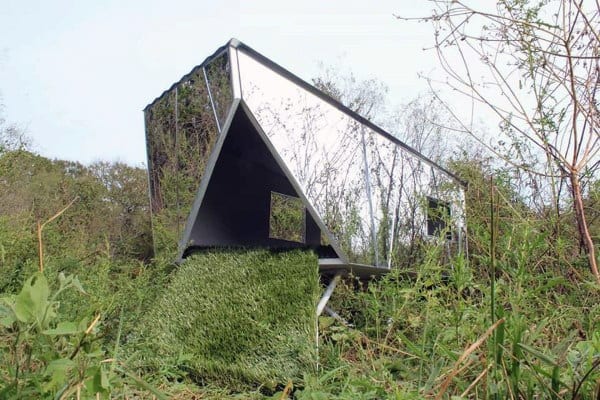 Unique dog house with a reflective mirrored surface and a grassy floor, blending into the surrounding nature with a modern, angular design