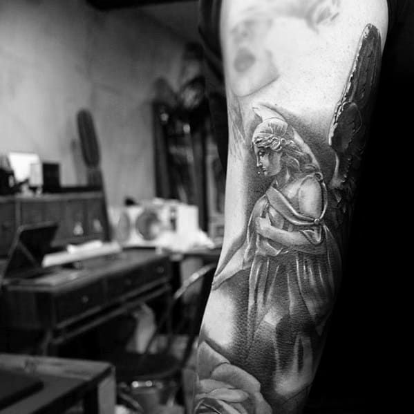 A black and grey tattoo on the forearm featuring an angel in a contemplative pose, with detailed wings and a flowing robe, set against a blurred background.