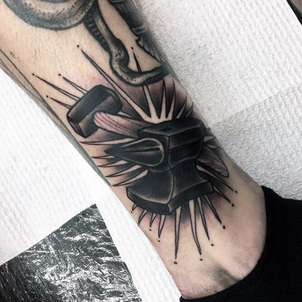Black and grey anvil tattoo on the leg with a hammer striking it, surrounded by bold radiating lines and shading for a dramatic, high-impact design.