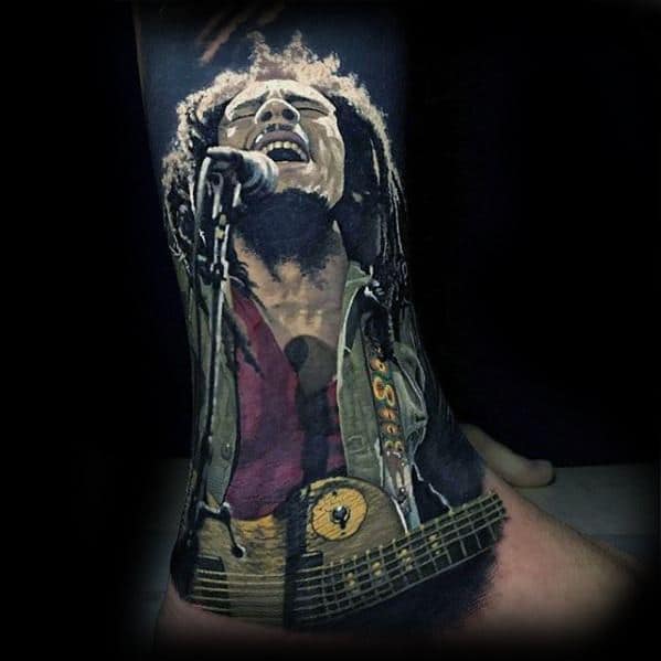 Lifelike color tattoo of Bob Marley singing passionately into a mic while playing guitar, capturing an energetic concert moment with vivid detail.
