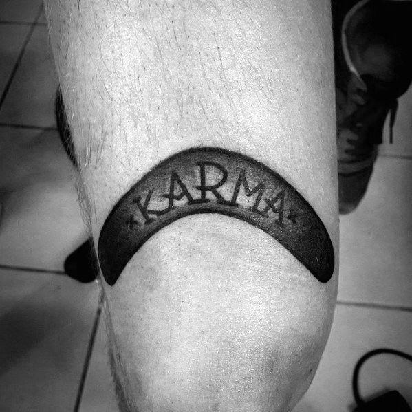 Solid black boomerang tattoo above the knee with the word KARMA and two stars etched in negative space at the center.