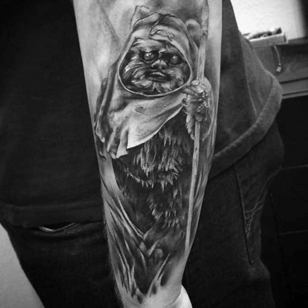Realistic black and grey Ewok tattoo on the forearm, featuring deep shading and dramatic lighting with a spear and intense expression.