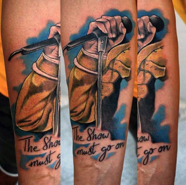 Color tattoo of Freddie Mercury's hand gripping a microphone in his yellow jacket, with the phrase “The Show Must Go On” below in script.