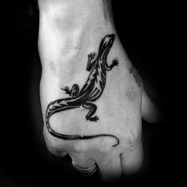 Black gecko tattoo on hand with a long curved tail and bold lightning-like patterns along the body.