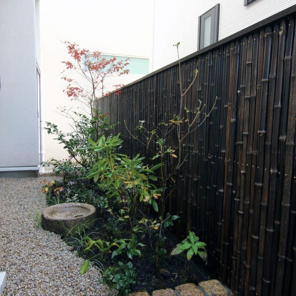 Dark bamboo privacy fence with small shrubs and plants along a gravel pathway in a narrow garden.
