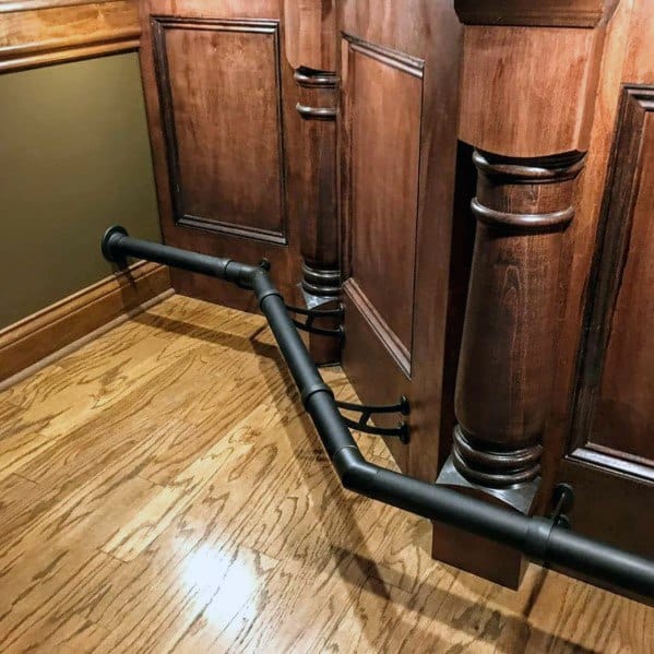 Matte black metal foot rail attached to a wooden bar with decorative columns on polished hardwood flooring.