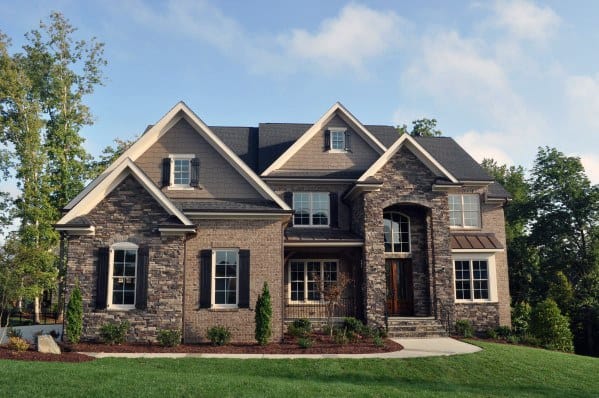 Elegant two-story home with a mix of dark brick and stone cladding, gabled rooflines, arched entryway, and a landscaped front yard