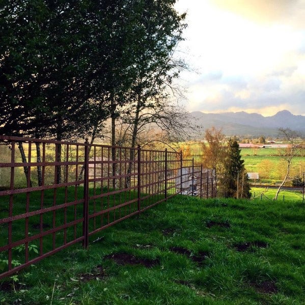 Rustic metal fence on a grassy hill, with trees and distant mountains under a partly cloudy sky