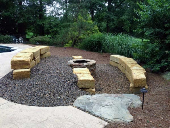 Curved stone bench and fire pit surrounded by trees and gravel in a backyard setting
