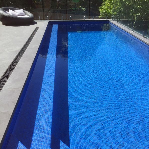 Modern pool with vibrant blue mosaic tiles, glass fencing, and a sleek concrete pool deck.