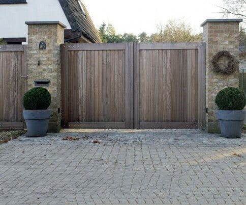 Wooden gate flanked by brick pillars, with round shrubs in large pots, on a paved driveway