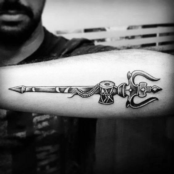 Forearm tattoo of Lord Shiva's trishul with a damru at the center, detailed with sharp lines and beads, symbolizing divine power and rhythm.