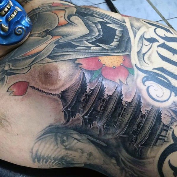 Chest tattoo featuring a black and grey Japanese pagoda with layered rooftops, surrounded by colorful flowers, script, and dramatic faces.