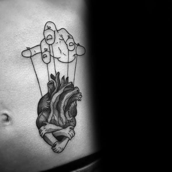 Bold linework puppet tattoo of a hand controlling an anatomical heart with arms hugging itself, symbolizing emotional control and self-protection.