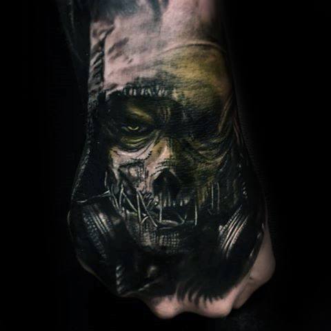 Dark scarecrow tattoo on the hand with a stitched mask, glowing yellow eye, gas mask, and eerie black and green shading.