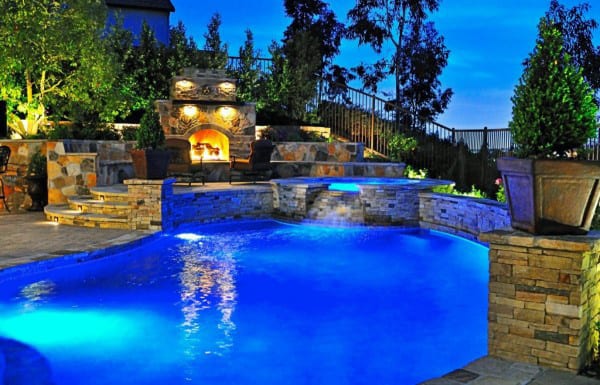 Illuminated backyard pool with stone walls and a lit outdoor fireplace, surrounded by trees under a twilight sky