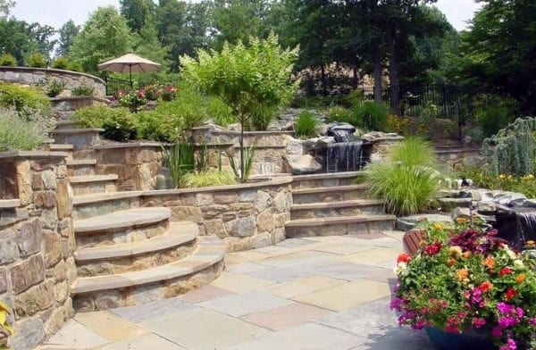 Multi-level stone retaining walls with stairs, waterfalls, lush greenery, and colorful flower planters.