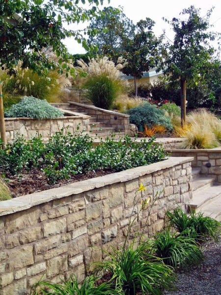 Layered stone retaining walls on a sloped landscape with plants, trees, and decorative grasses.