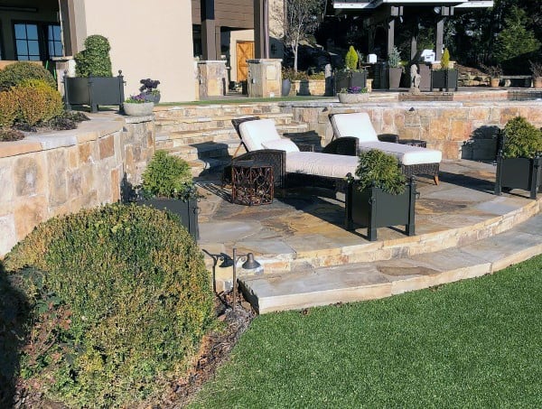 Stone retaining wall with patio, lounge chairs, potted plants, and landscaped greenery.