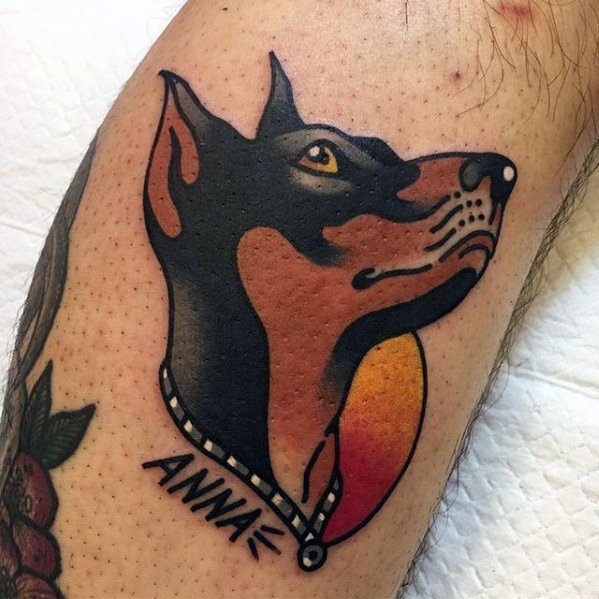 Traditional Doberman tattoo with clean black outlines, sunset background, and a name tag reading “Anna” for a bold, personal tribute.