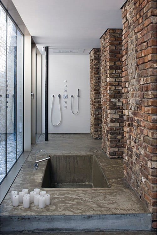 Concrete bathtub with exposed brick columns, glass wall, and minimalist shower design.