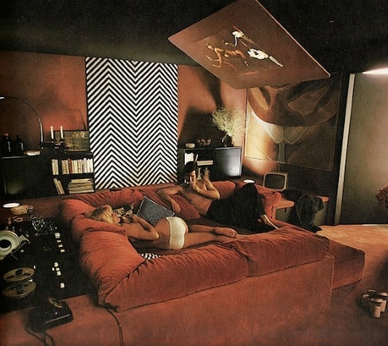 People lounging on a red sectional sofa in a retro room with patterned walls and vintage electronics