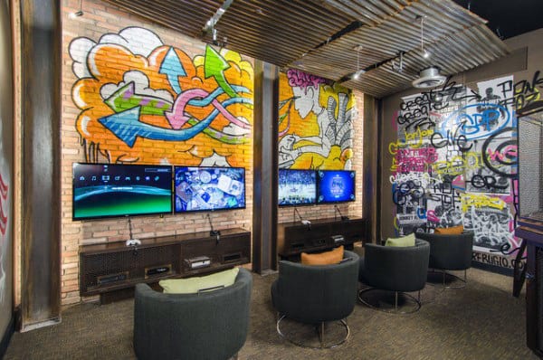 Gaming room with graffiti walls, three TVs mounted on brick, and round chairs arranged in a cozy setup