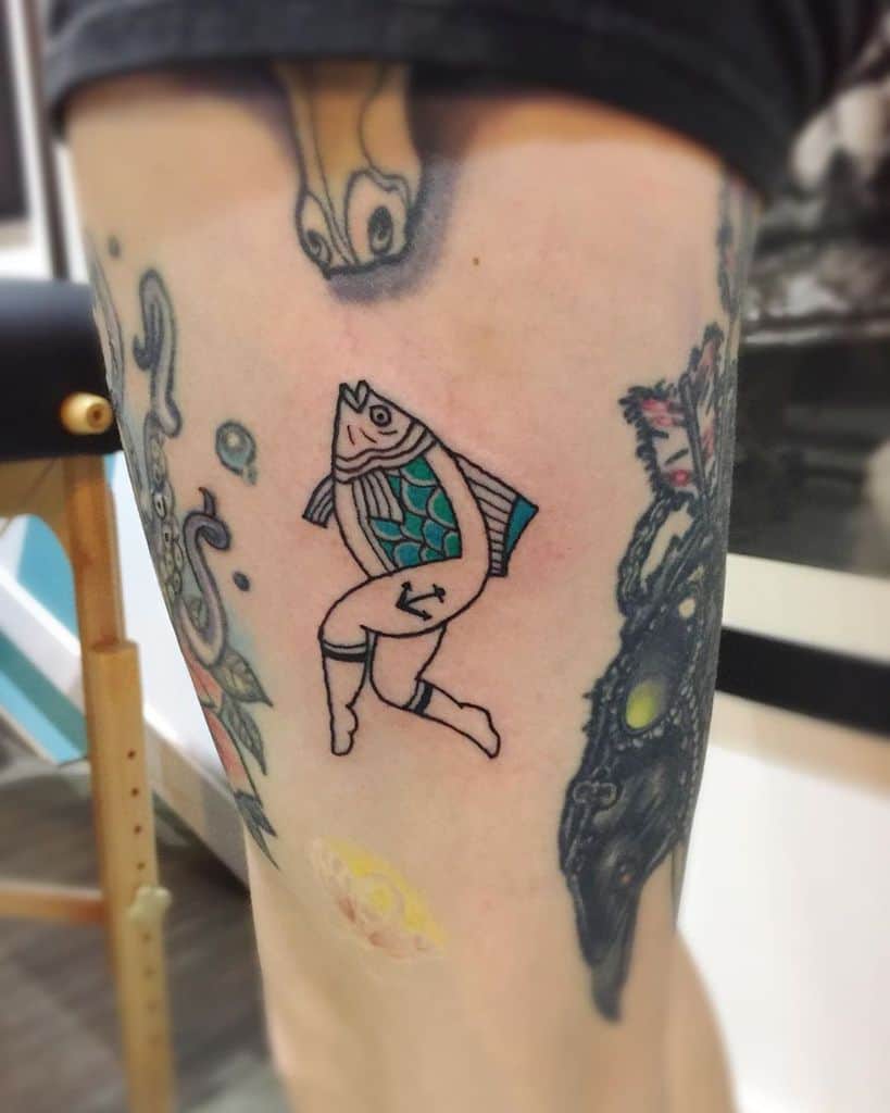 A colorful tattoo of a fish with human legs wearing socks and garters, drawn in a whimsical and humorous style on the thigh.