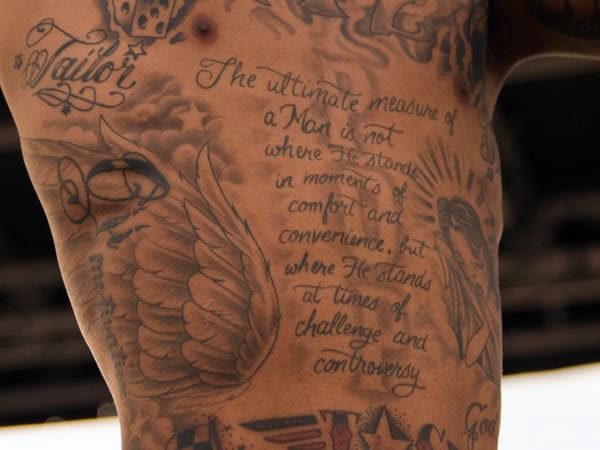 Close-up of various tattoos on torso, featuring text and angel wings