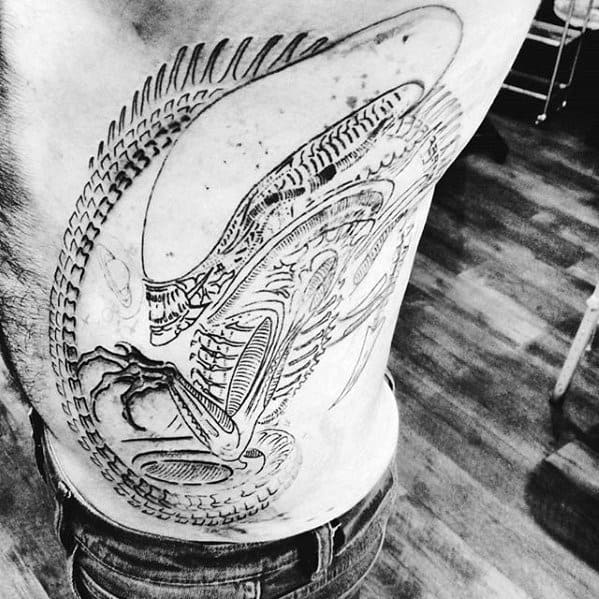 Large black and white Xenomorph tattoo across the side torso, featuring the alien in a curled pose with detailed skeletal and tail linework.