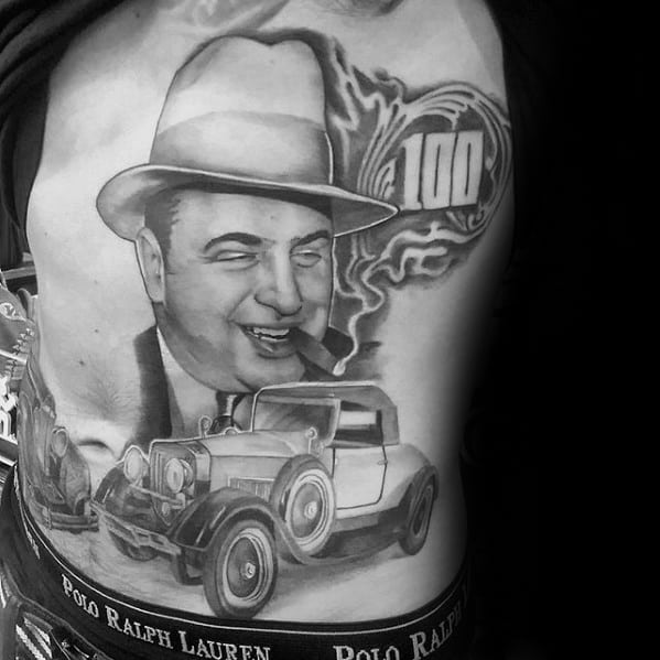 Al Capone tattoo on the chest featuring a black-and-white portrait of the gangster with a cigar, alongside a vintage car and the number "100," capturing his iconic gangster image and era.