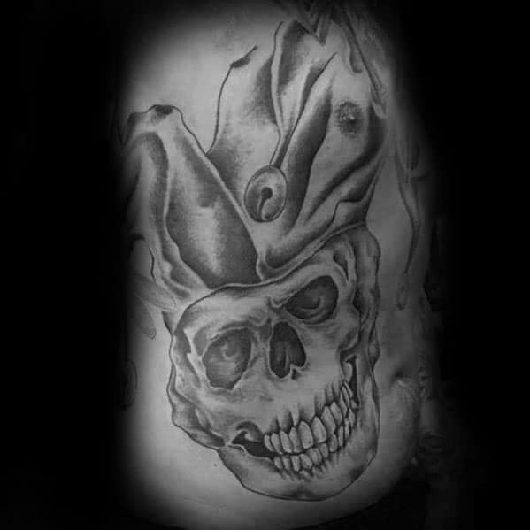 A black and gray tattoo of a skull with a jester hat, featuring intricate shading and details, placed on the side of the body.