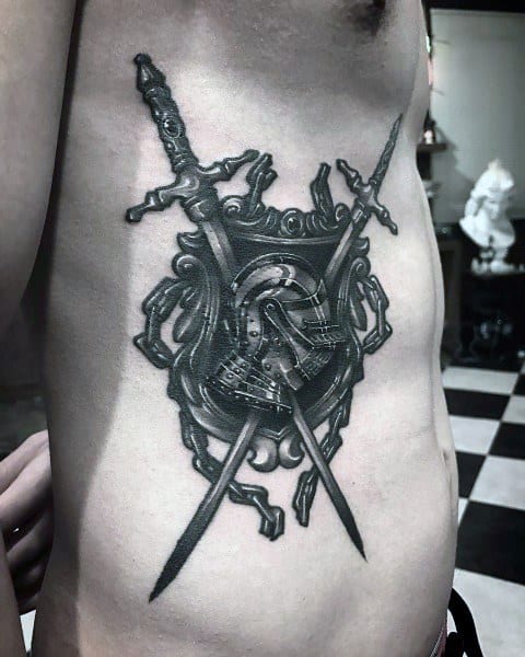 Side torso tattoo of a knight’s helmet within an ornate shield, flanked by crossed swords, done in bold black and grey shading.