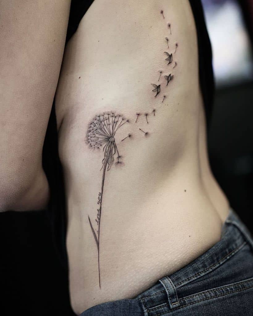 A side rib tattoo of a dandelion with seeds blowing off and a light trail in the background, symbolizing freedom and movement.
