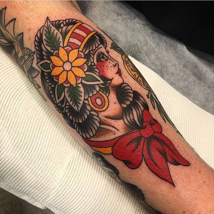 Traditional tattoo of a gypsy woman with a floral headpiece, bold shading, and a large red bow on the forearm.