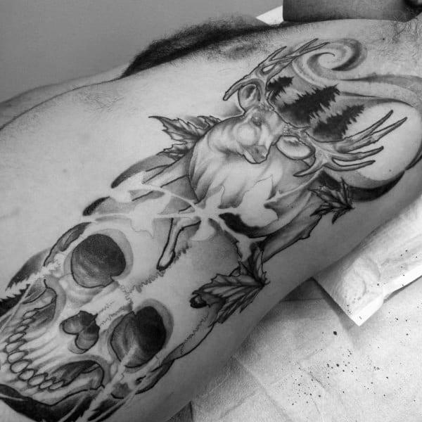 Black and grey tattoo featuring a realistic buck with antlers transitioning into forest trees above a cracked human skull and maple leaves.