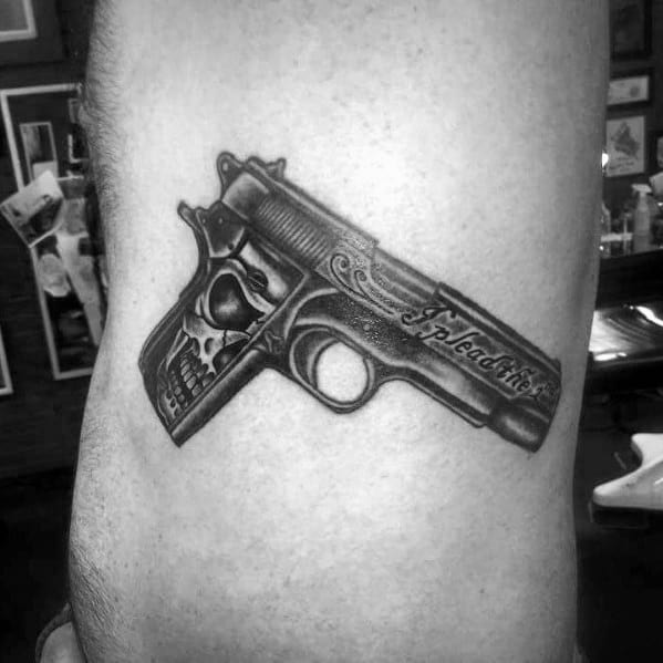 Realistic black and gray tattoo of an engraved 1911 pistol with a skull design on the grip and intricate detailing, placed on the side of the torso.