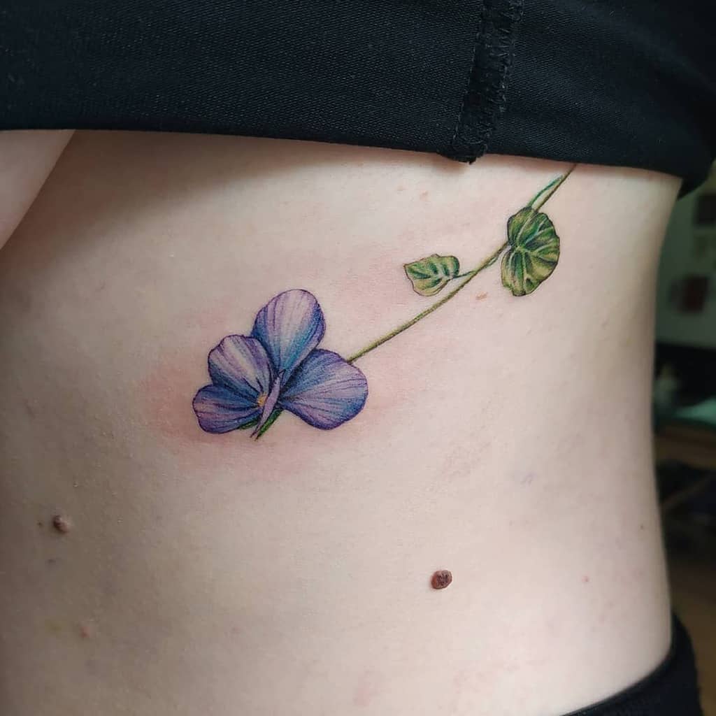 Minimalist rib tattoo of a delicate violet flower with a trailing stem and leaves, symbolizing elegance, growth, and simplicity