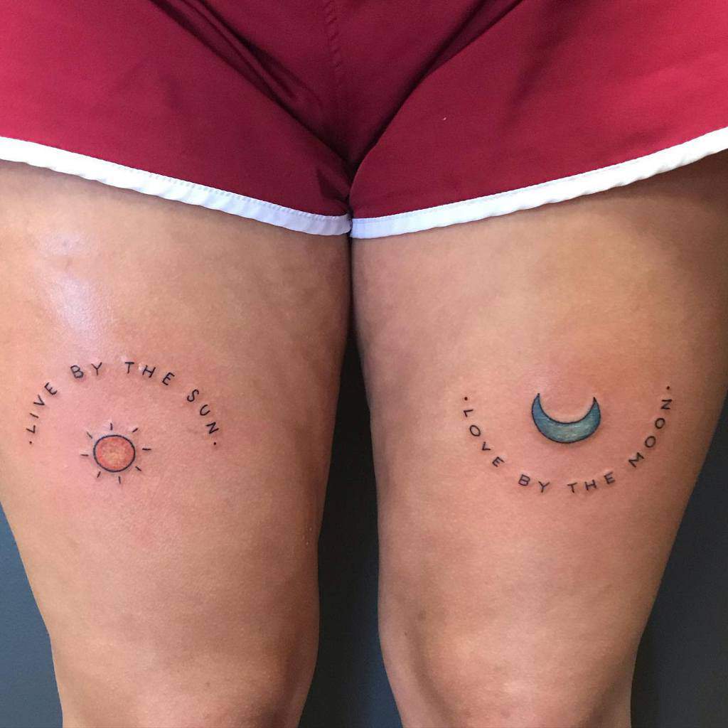 Matching thigh tattoos featuring a small sun with "Live by the Sun" on one leg and a crescent moon with "Love by the Moon" on the other.