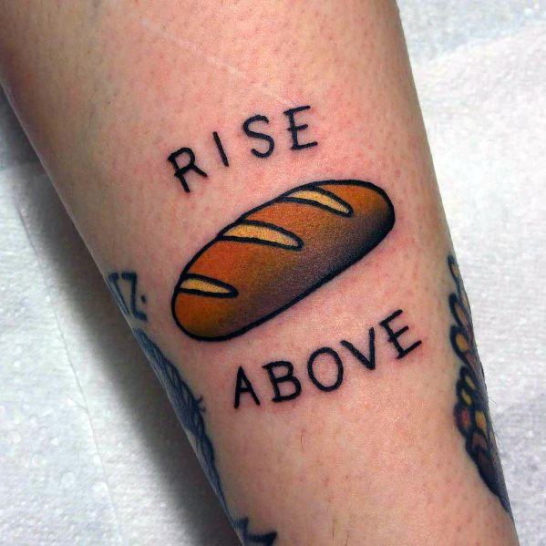 Colorful tattoo of a golden brown loaf of bread with the phrase "Rise Above," blending a playful pun with bold traditional-style lettering.