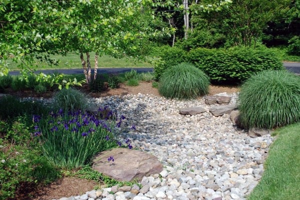 Peaceful garden with a mix of river rocks, purple flowers, and lush green plants, creating a natural and relaxing landscape