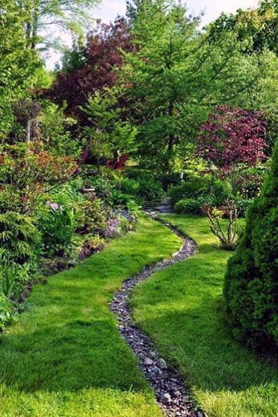 Charming garden with a winding river rock path through lush green grass, surrounded by vibrant plants and trees, creating a peaceful atmosphere