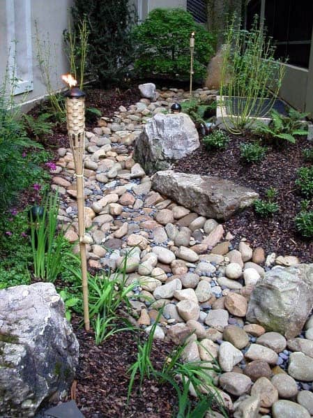 River rock path with large stones and tiki torches, surrounded by lush greenery and mulch, creating a tranquil outdoor walkway